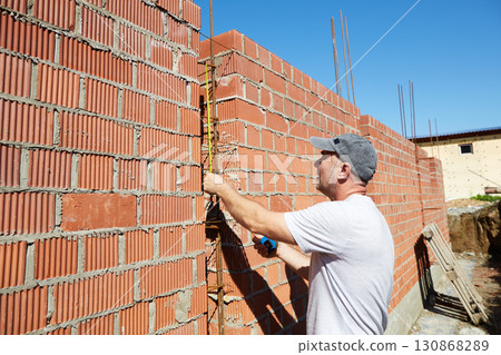 A construction worker is measuring a brick wall at a building site A construction worker is measuring a brick wall at a building site 130868289