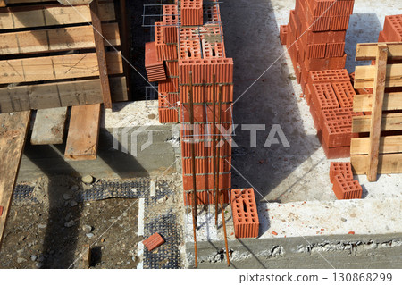 Workers are constructing walls with red bricks at a residential site, with wooden scaffolding and freshly laid foundation visible Workers are constructing walls with red bricks at a residential site, with wooden scaffolding and freshly laid foundation visible 130868299