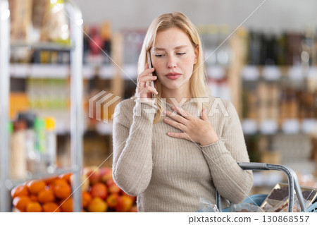 Upset young woman choosing groceries while talking on phone Upset young woman choosing groceries while talking on phone 130868557