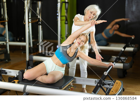 Senior woman trainer helps young woman to perform exercises in sitting position on pilates reformer Senior woman trainer helps young woman to perform exercises in sitting position on pilates reformer 130868584