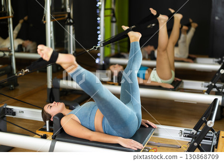 Woman lying on reformer and pulling adjustable cables with her legs 130868709