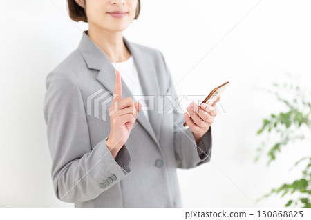 A middle-aged female business woman in a suit holding a smartphone and pointing her index finger up 130868825