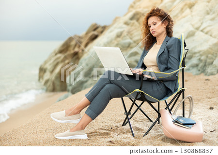 Pretty woman in office clothes, in suit, sitting on chair and working on laptop on seashore Pretty woman in office clothes, in suit, sitting on chair and working on laptop on seashore 130868837