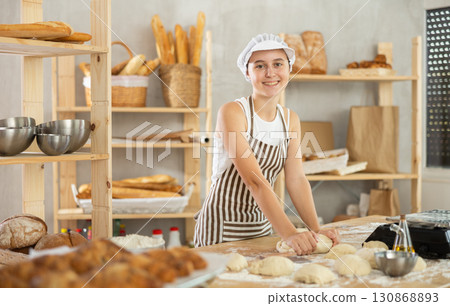 Teen girl works in bakery as baker, kneads dough, works with flour. Teen girl works in bakery as baker, kneads dough, works with flour. 130868893