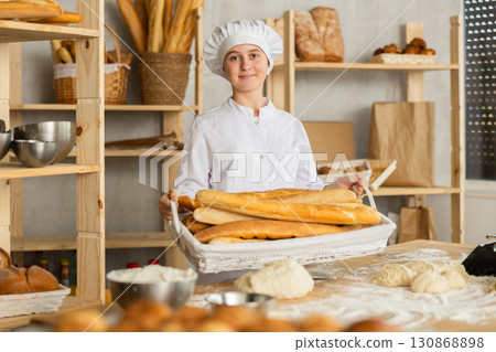 Young girl baker are standing near product range in bakery kitchen with basket loaf of baguettes Young girl baker are standing near product range in bakery kitchen with basket loaf of baguettes 130868898