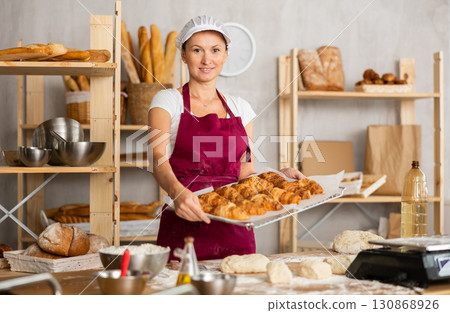 Positive female baker in uniform stands behind counter in private bakery offering croissants 130868926