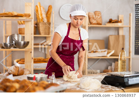 Smiling young female professional baker in apron standing at work table and kneading dough during working day in bakery 130868937