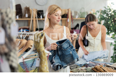 Fashionista girl buying denim shorts. Female client choosing something in background Fashionista girl buying denim shorts. Female client choosing something in background 130869009