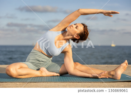 Female fitness trainer performing yoga parsvottanasana sitting on mat on beach 130869061