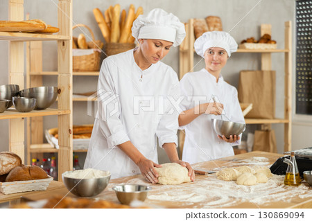 Two professional female baker in white uniform working in bakehouse, whips up and kneads dough 130869094