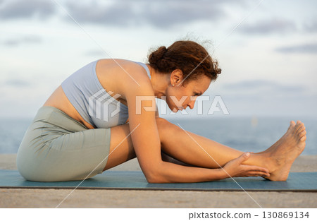 Yoga practice on embankment, female athlete fulfil carry out Pashchimottanasana 130869134