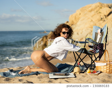 Naked woman in open shirt kneeling by chair on sandy seashore Naked woman in open shirt kneeling by chair on sandy seashore 130869183