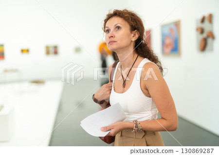 Interested woman examining paintings on walls in art gallery Interested woman examining paintings on walls in art gallery 130869287