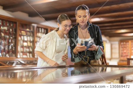 Woman with smartphone and teenage girl looking at exhibit in museum display Woman with smartphone and teenage girl looking at exhibit in museum display 130869472