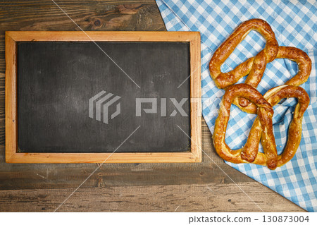 Top view of blackboard with pretzel loaf on a blue checkered tablecloth or napkin with a wooden table background. Copy space. Concept of Oktoberfest. Bavarian German culture festival decoration symbol Top view of blackboard with pretzel loaf on a blue checkered tablecloth or napkin with a wooden table background. Copy space. Concept of Oktoberfest. Bavarian German culture festival decoration symbol 130873004