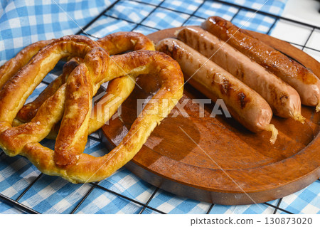 Pretzel loaf and sausage on a wooden tray with a blue checkered tablecloth or napkin background. Concept of Oktoberfest. Bavarian German culture festival decoration symbol Pretzel loaf and sausage on a wooden tray with a blue checkered tablecloth or napkin background. Concept of Oktoberfest. Bavarian German culture festival decoration symbol 130873020