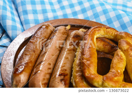 Pretzel loaf and sausage on a wooden tray with a blue checkered tablecloth or napkin background. Concept of Oktoberfest. Bavarian German culture festival decoration symbol Pretzel loaf and sausage on a wooden tray with a blue checkered tablecloth or napkin background. Concept of Oktoberfest. Bavarian German culture festival decoration symbol 130873022