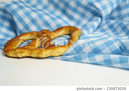 Closeup view of a pretzel loaf with a blue checkered tablecloth or napkin in a white table background. Concept of Oktoberfest. Bavarian German culture festival decoration symbol Closeup view of a pretzel loaf with a blue checkered tablecloth or napkin in a white table background. Concept of Oktoberfest. Bavarian German culture festival decoration symbol 130873039