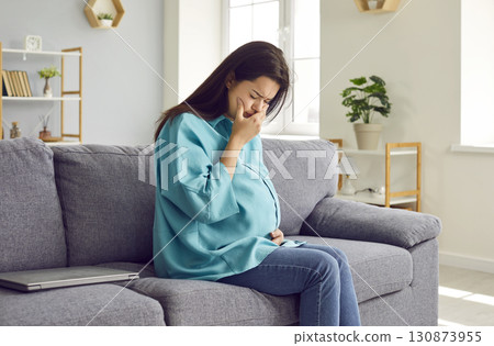 Young pregnant woman sitting on couch, having nausea and holding hand on her mouth Young pregnant woman sitting on couch, having nausea and holding hand on her mouth 130873955