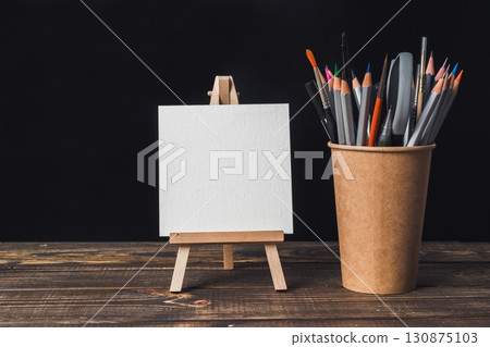 White canvas is on a wooden table next to a cup of colored pencils White canvas is on a wooden table next to a cup of colored pencils 130875103