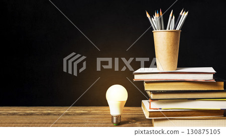 Light bulb is lit up on a table with a stack of books and pencils 130875105
