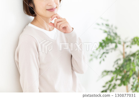 A middle-aged woman in casual clothes leaning against a white wall by the window 130877031