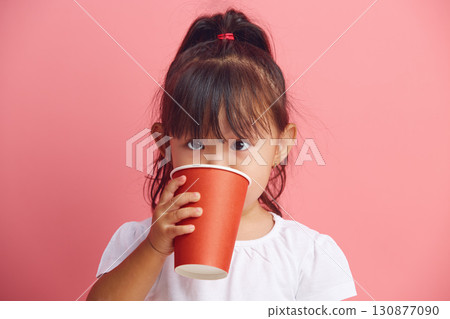 Funny little Asian girl holds disposable paper cup in her hands and drinks water standing on a pink isolated background.  130877090