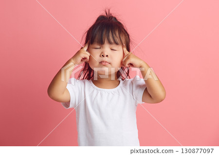 Serious little asian ethnicity girl keeps fingers on temples and closes eyes to remember something important standing isolated over pink background. Serious little asian ethnicity girl keeps fingers on temples and closes eyes to remember something important standing isolated over pink background. 130877097