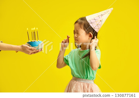 Chinese Little Kid Girl Wishing on Birthday Candles Stands Against Yellow Isolated Background, Closing her Eyes and Crossing Fingers, Wearing in Party Hat, Expressing Hope for Wish Fulfillment  130877161