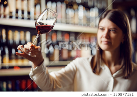 Profesional female bartender rates red wine, looks at its color, evaluates the density and tartness of an alcoholic drink, stands in cellar on shelf with different wines background. 130877181