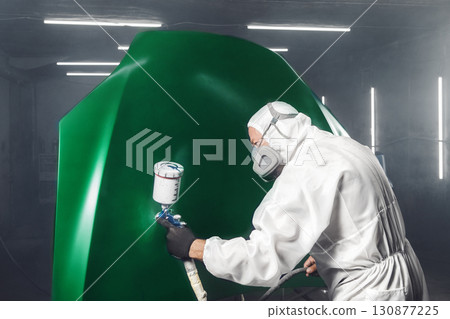 Worker paints car hood to green color in workshop chamber using airbrush. Man using spray gun during auto painting. Worker paints car hood to green color in workshop chamber using airbrush. Man using spray gun during auto painting. 130877225