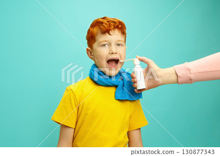 Seasonal Colds in Children Concept. Little Red Headed Boy Wrapped in a Scarf Preparing to Take Cough Throat Spray From Hands of Adult Standing on Blue Isolated Background Seasonal Colds in Children Concept. Little Red Headed Boy Wrapped in a Scarf Preparing to Take Cough Throat Spray From Hands of Adult Standing on Blue Isolated Background 130877343