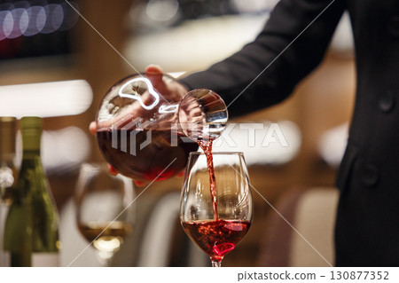 Professional female sommelier pours red wine from decanter to the glass, close up image. Woman waiter pouring alcoholic drink being in cellar of wine shop. 130877352
