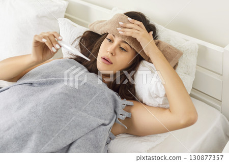 Unhealthy woman with high temperature measures with thermometer, feels bad, sick ill suffers  130877357