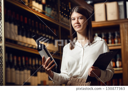 Professional caucasian woman sommelier in wine cellar making notes in tablet and choosing wine from shelve. Association of professional winemaker. Professional caucasian woman sommelier in wine cellar making notes in tablet and choosing wine from shelve. Association of professional winemaker. 130877363
