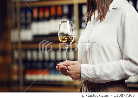 Vintner woman tasting white wine from a glass in cellar. Close up shot of female sommelier hand that hold the wine glass on shelf background. Vintner woman tasting white wine from a glass in cellar. Close up shot of female sommelier hand that hold the wine glass on shelf background. 130877366