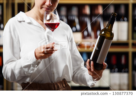 Young beautiful woman sommelier tasting red wine in the wine cellar, close up photo. Alcoholic beverages degustation. Winemaking school, wine waiter certification. 130877370