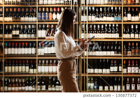 Female expert sommelier locating in winery sphere analyses various varieties of red and white wine, located in wine cellar, making notes in tablet.  130877372