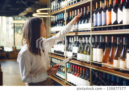 Professional woman sommelier getting wine bottle from shelves, making notes on tablet being at wine cellar. 130877377