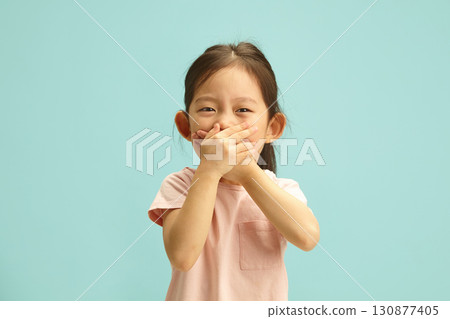 Cheerful Asian ethnicity Kid Girl with Hand Over Mouth, does not want to tell the truth, can barely contain her laughter, tries to deceive or keep silent about something standing over blue isolated. Cheerful Asian ethnicity Kid Girl with Hand Over Mouth, does not want to tell the truth, can barely contain her laughter, tries to deceive or keep silent about something standing over blue isolated. 130877405