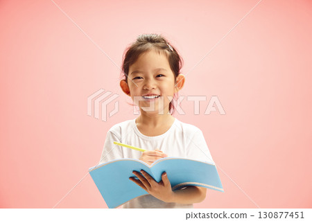 Cheerful First Grader Chinese Girl Smiling while Holding Exercise Book and Pen Standing on Pink Isolated Cheerful First Grader Chinese Girl Smiling while Holding Exercise Book and Pen Standing on Pink Isolated 130877451