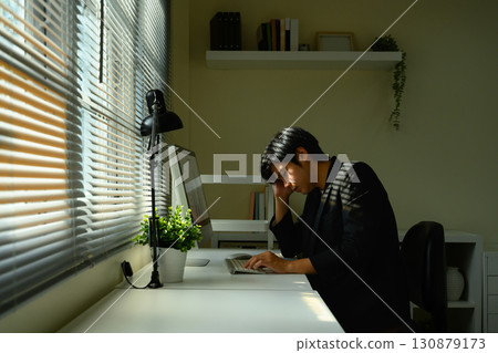 Stressed businessman sitting at desk with hand on head while working on computer in dimly lit office 130879173