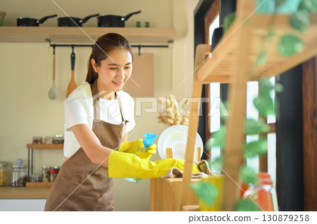 Smiling young woman in apron and rubber gloves cleaning kitchen counter with spray bottle and cloth 130879258