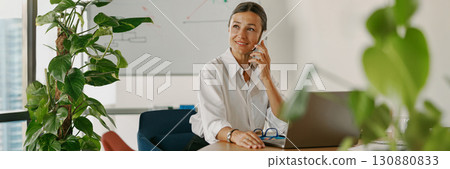 A professional businesswoman is working in a modern office environment while making a phone call 130880833