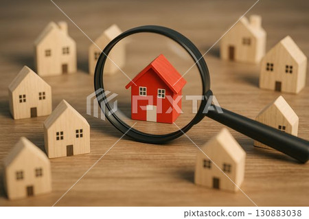 Red house under magnifying glass surrounded by wooden model homes on wooden surface 130883038