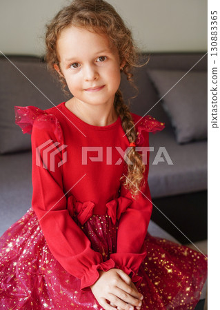 A young girl sits indoors wearing a red festive dress , smiling gently at the camera 130883365