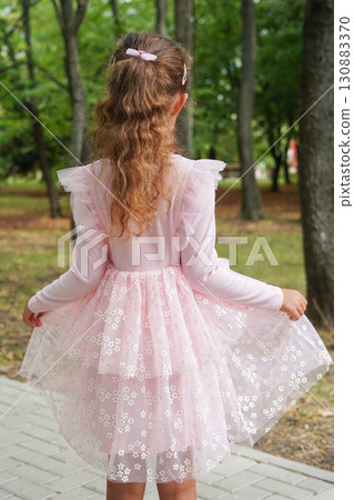 A young girl with curly hair and a bow clip is seen from behind wearing a layered pink floral dress 130883370