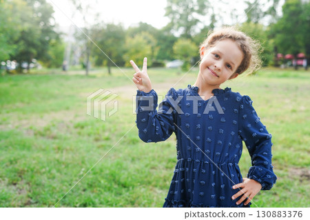 A smiling young girl wearing a navy embroidered dress poses outdoors with a playful peace sign A smiling young girl wearing a navy embroidered dress poses outdoors with a playful peace sign 130883376