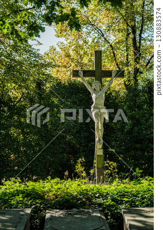 White statue of a crucifix surrounded by lush greenery in a serene outdoor setting 130883574