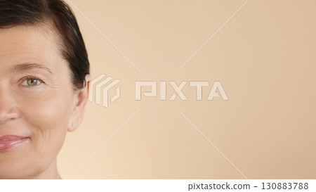 Close-up of happy female face on beige background, free space for advertising cosmetics, beauty care, skin care, spa salons. Half-face of a beautiful Caucasian middle-aged woman 50s age. 130883788